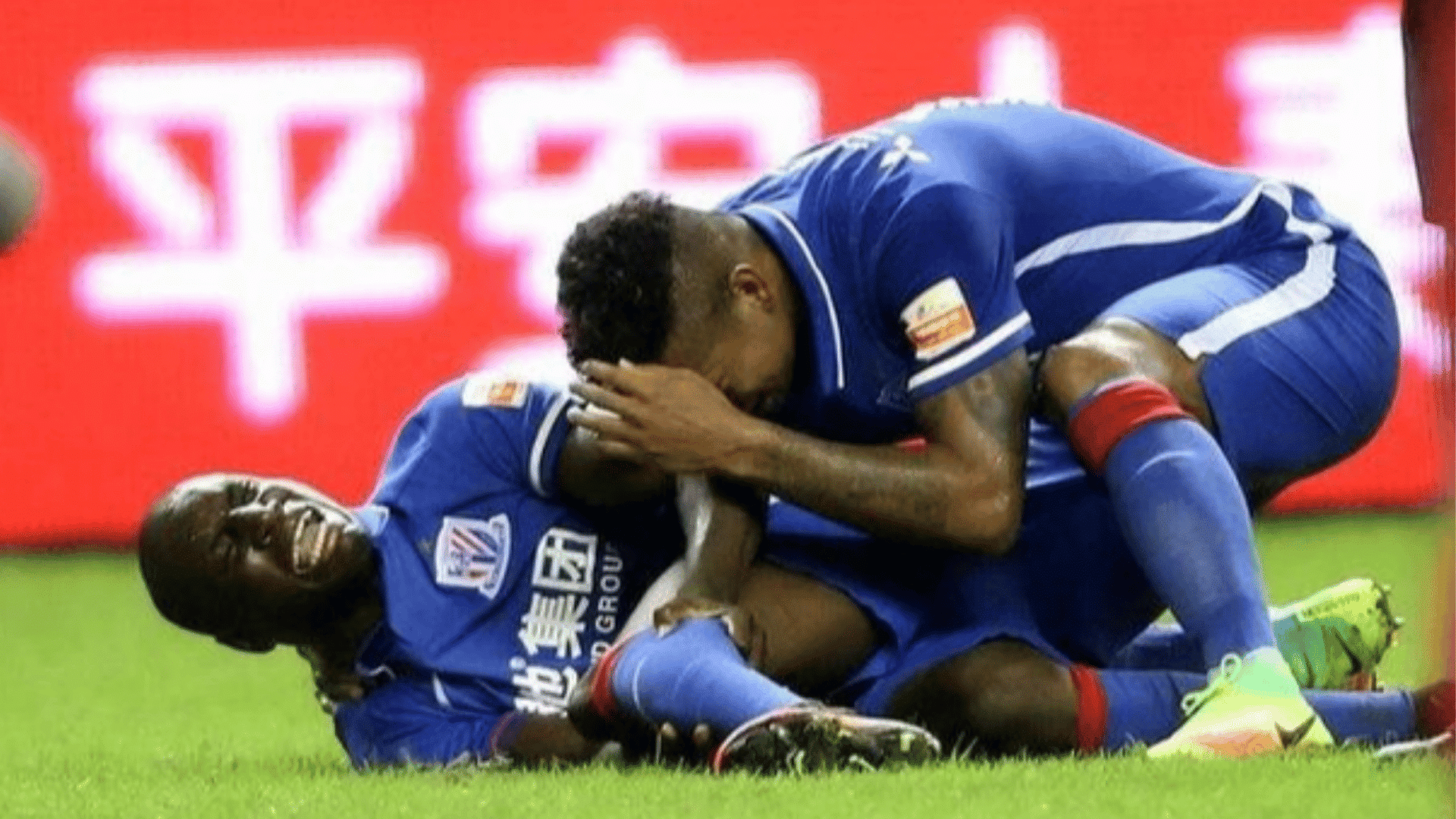 two soccer players in blue uniforms celebrate on the grass with one lying down smiling and another leaning over him