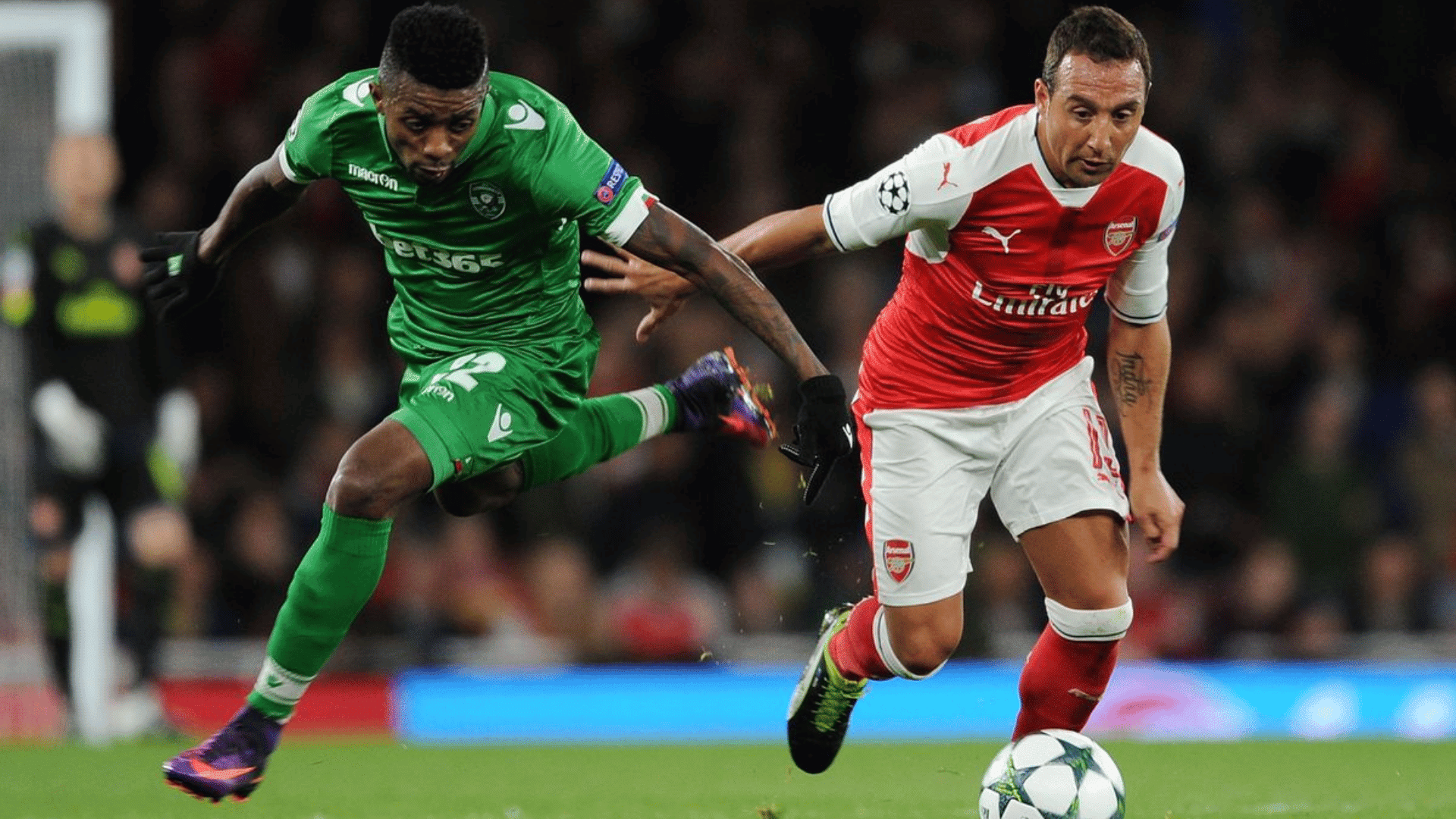 two soccer players race for the ball as one in green leaps beside another in red and white during a match