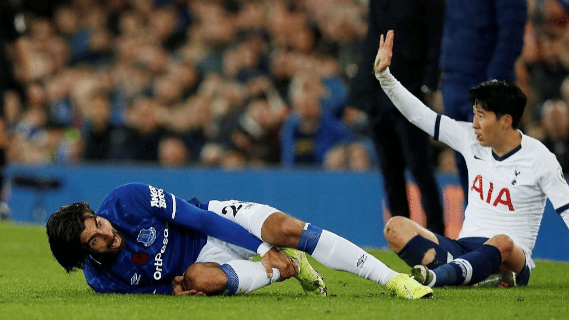 two soccer players sit on the grass after a tackle as one holds his leg and the other raises his hand