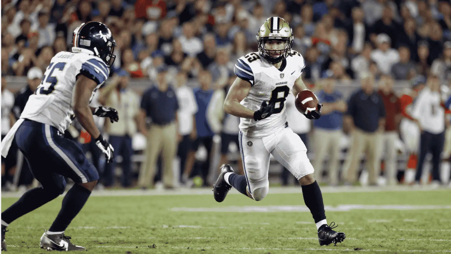 football player carries the ball across the field while a defender closes in during a night game