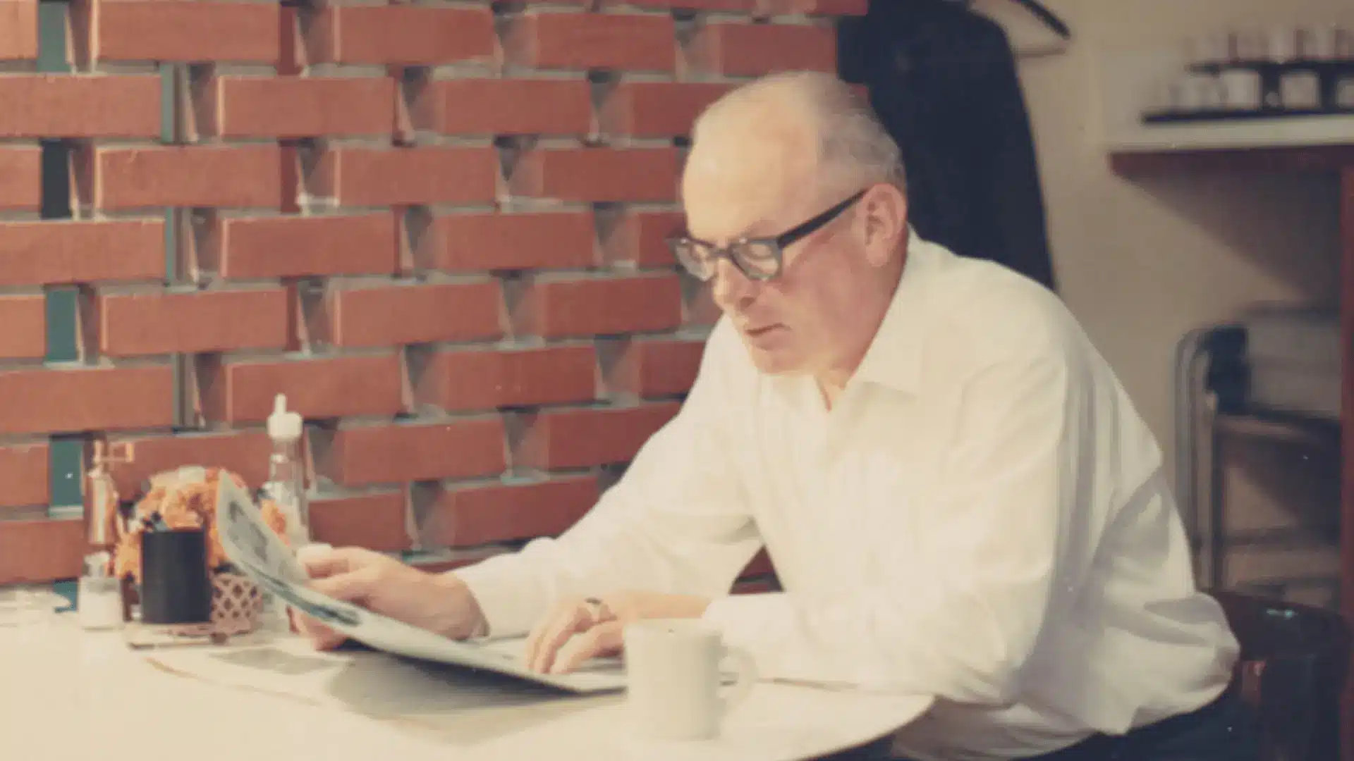image of bill winkenbach in a desk looking over at a newspaper with a red brick wall at the back