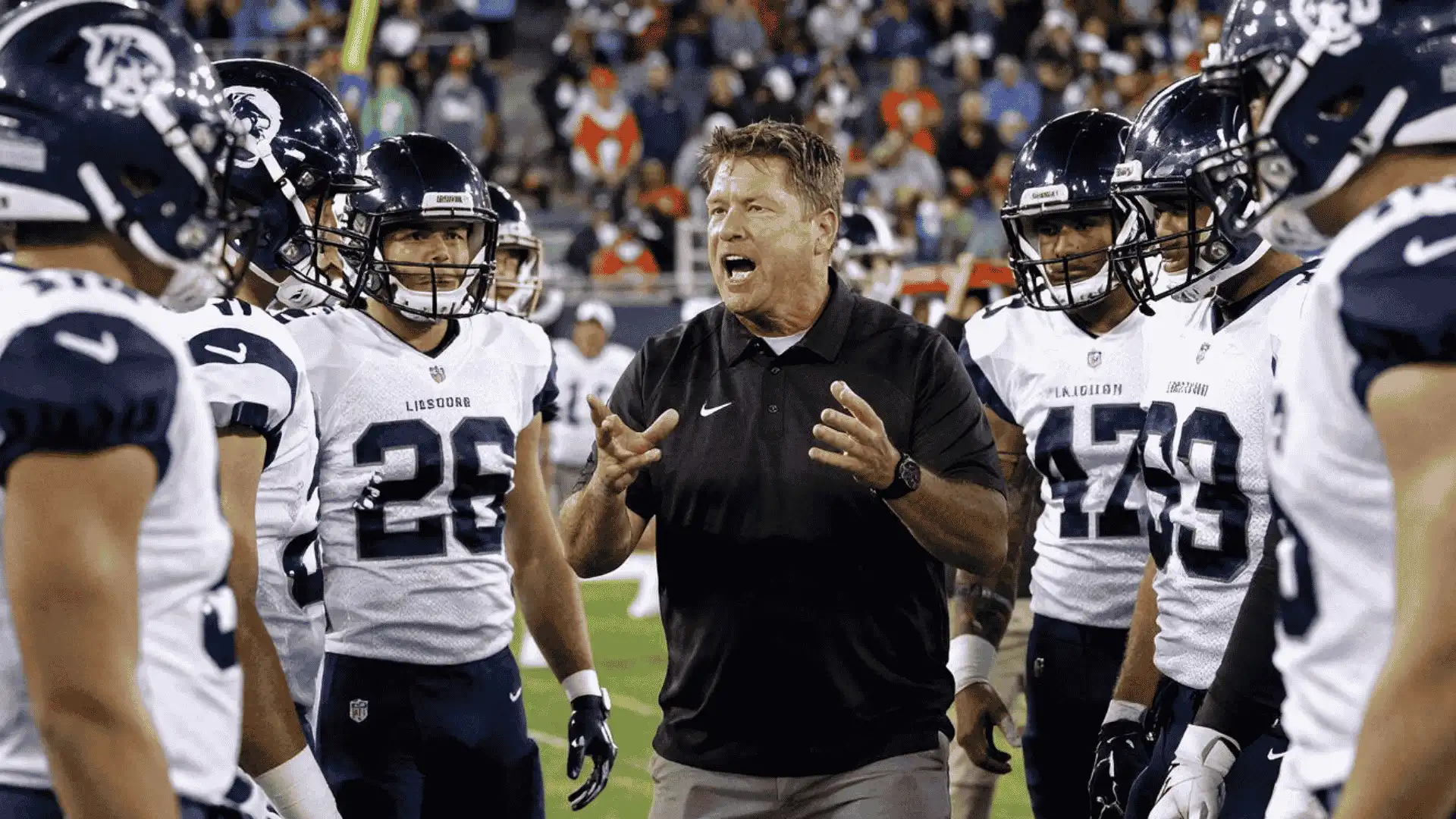 coach speaks to football players gathered in a sideline huddle during a crowded night game