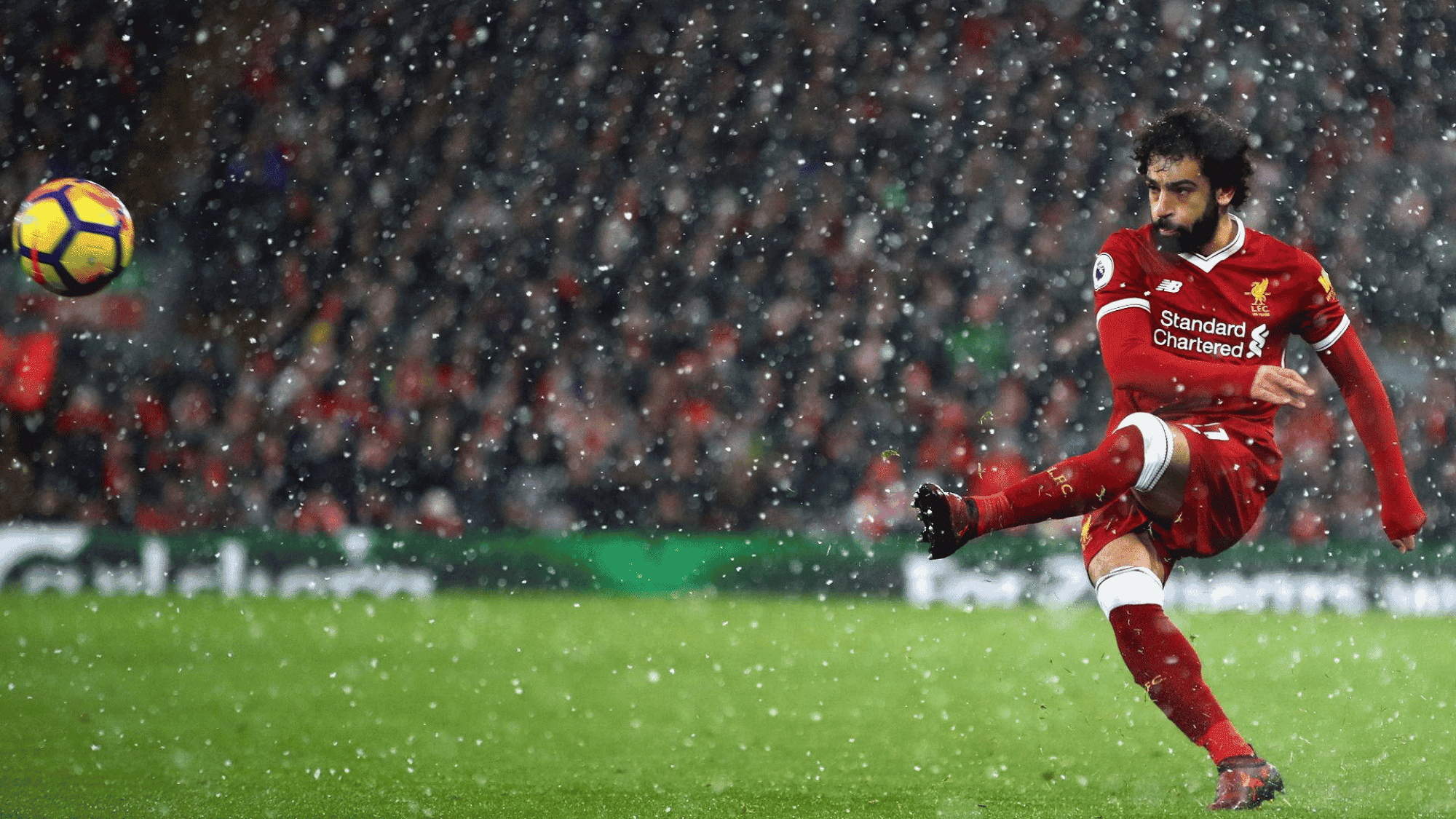wide angle focus shot on mohammed salah shooting the ball towards the goal on a rainy day wearing his red liverpool jersey