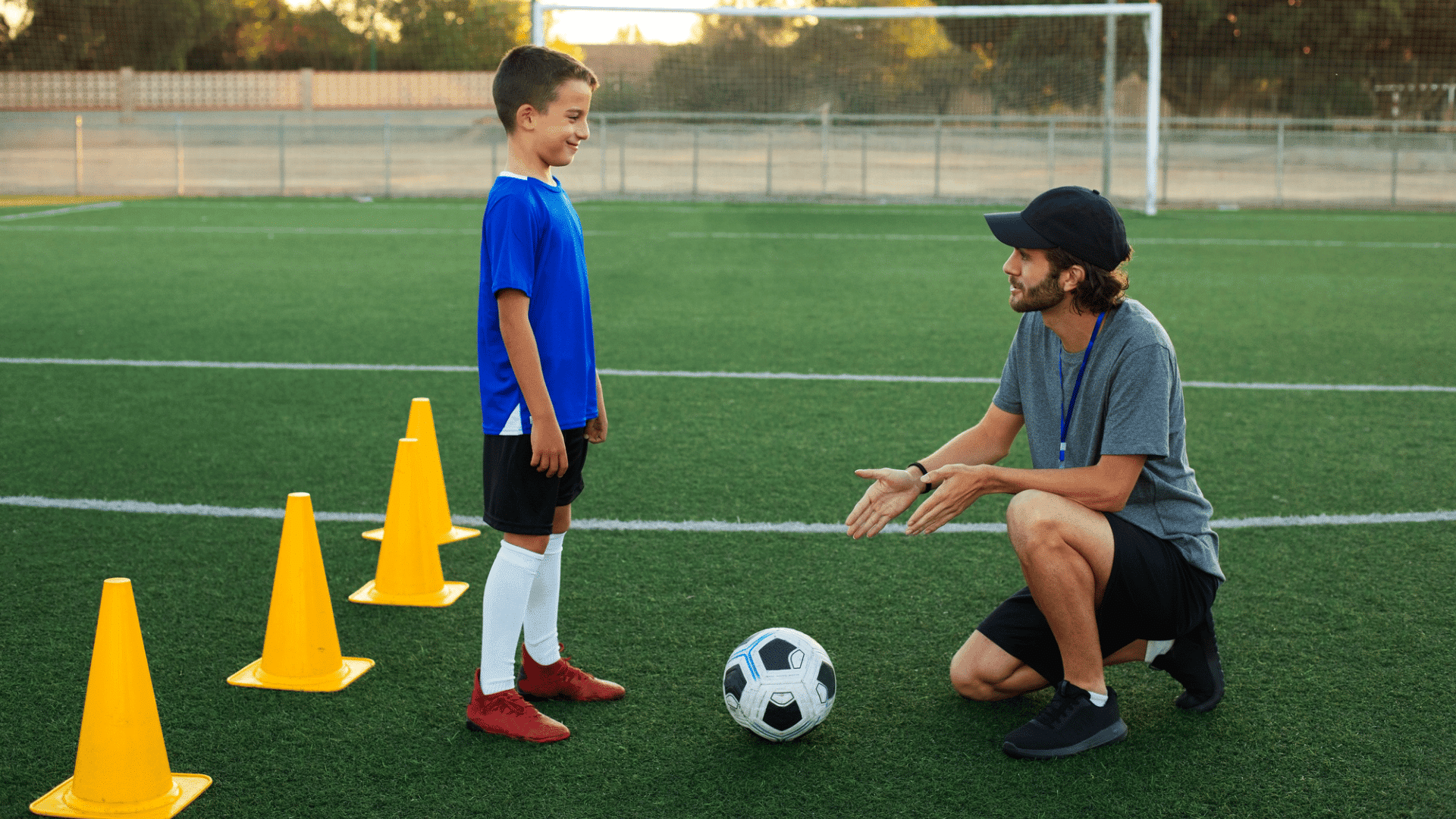 young boy in blue jersey listening closely to his kneeling coach on an outdoor training pitch with yellow marker cones