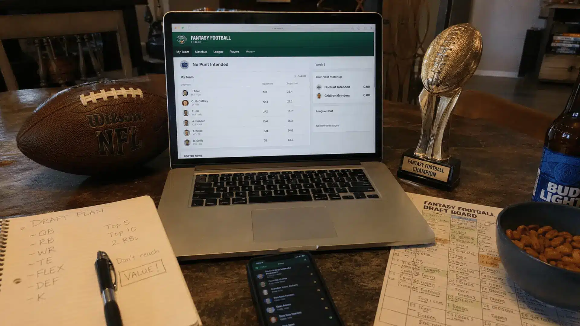 a laptop showing a fantasy football roster beside a football trophy draft notes phone snacks and a beer