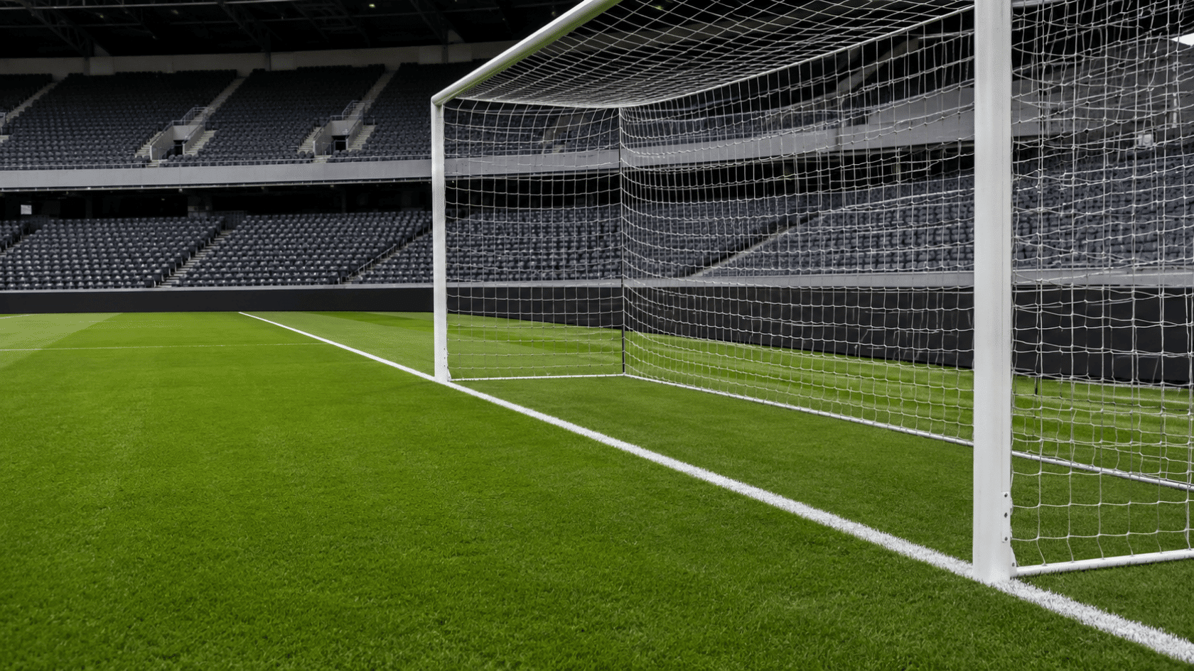 a white field marking runs beside the soccer goal across green grass with empty stadium seats behind