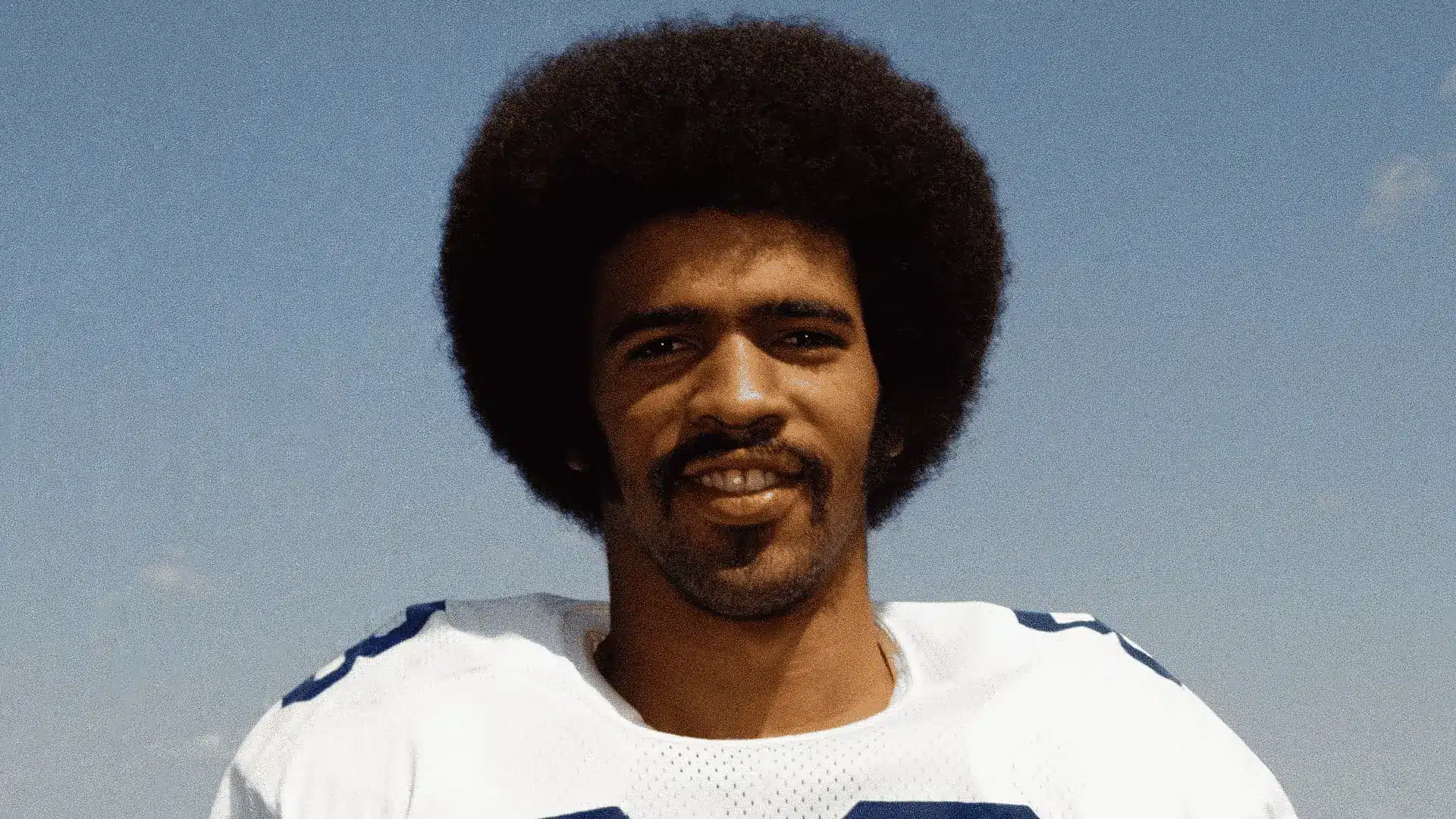 football player poses outdoors in a white jersey under a clear blue sky with calm expression
