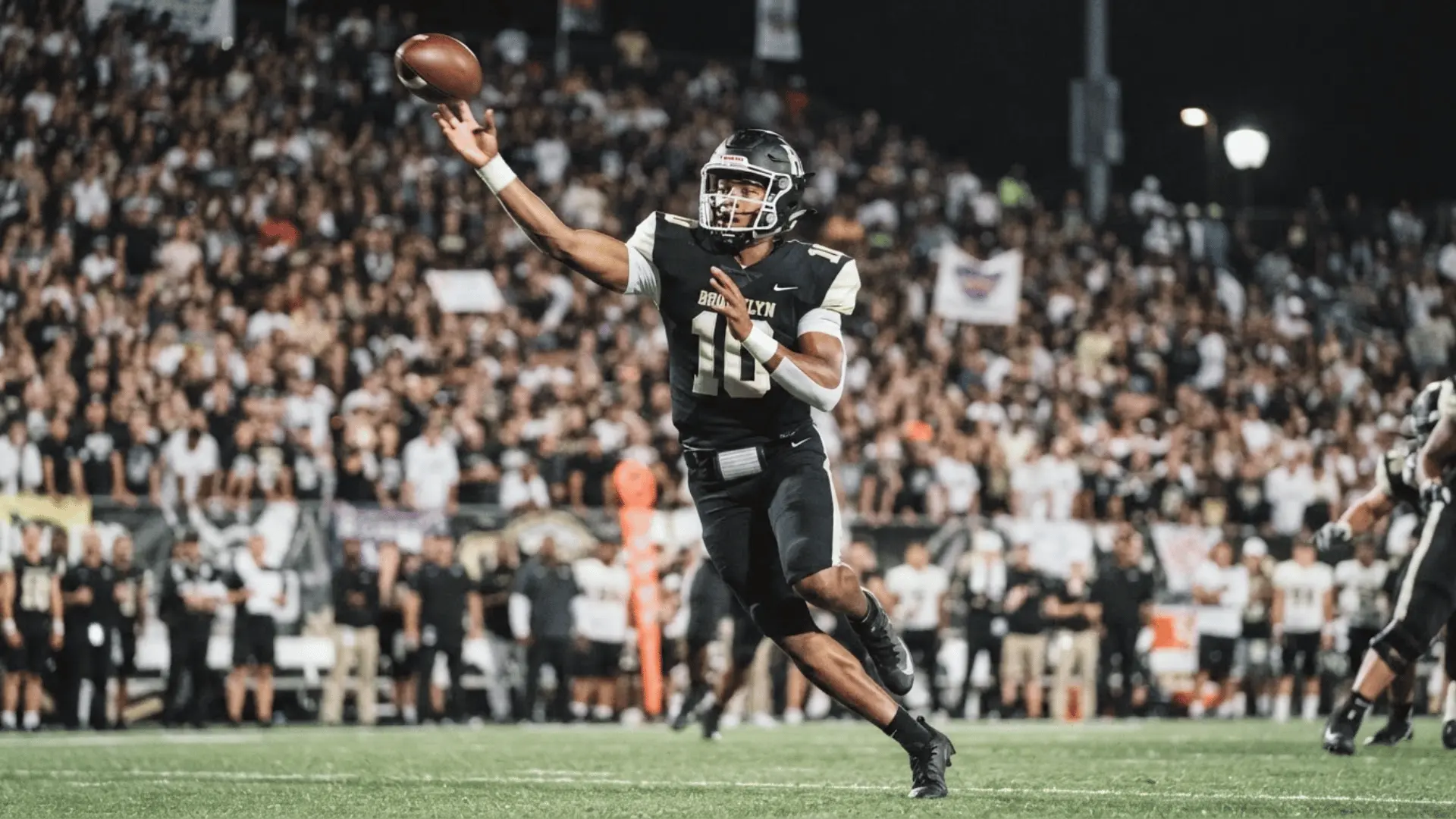football quarterback in black uniform throwing a pass during a night game in front of a crowded stadium