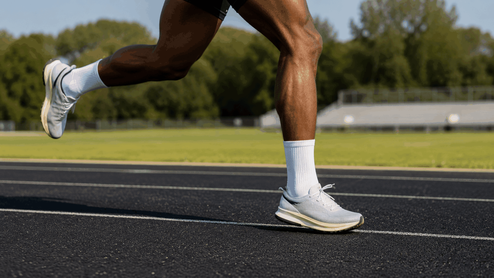 lower legs of a runner in white shoes on a track with grass and stadium in background