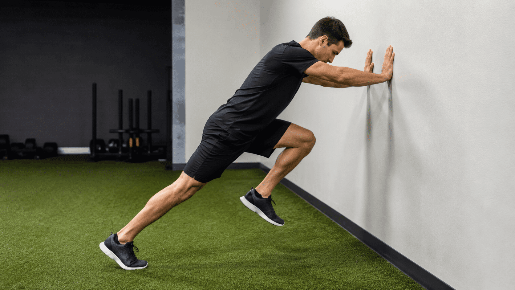 man in black athletic wear performing a wall sprint drill in a gym leaning against a wall
