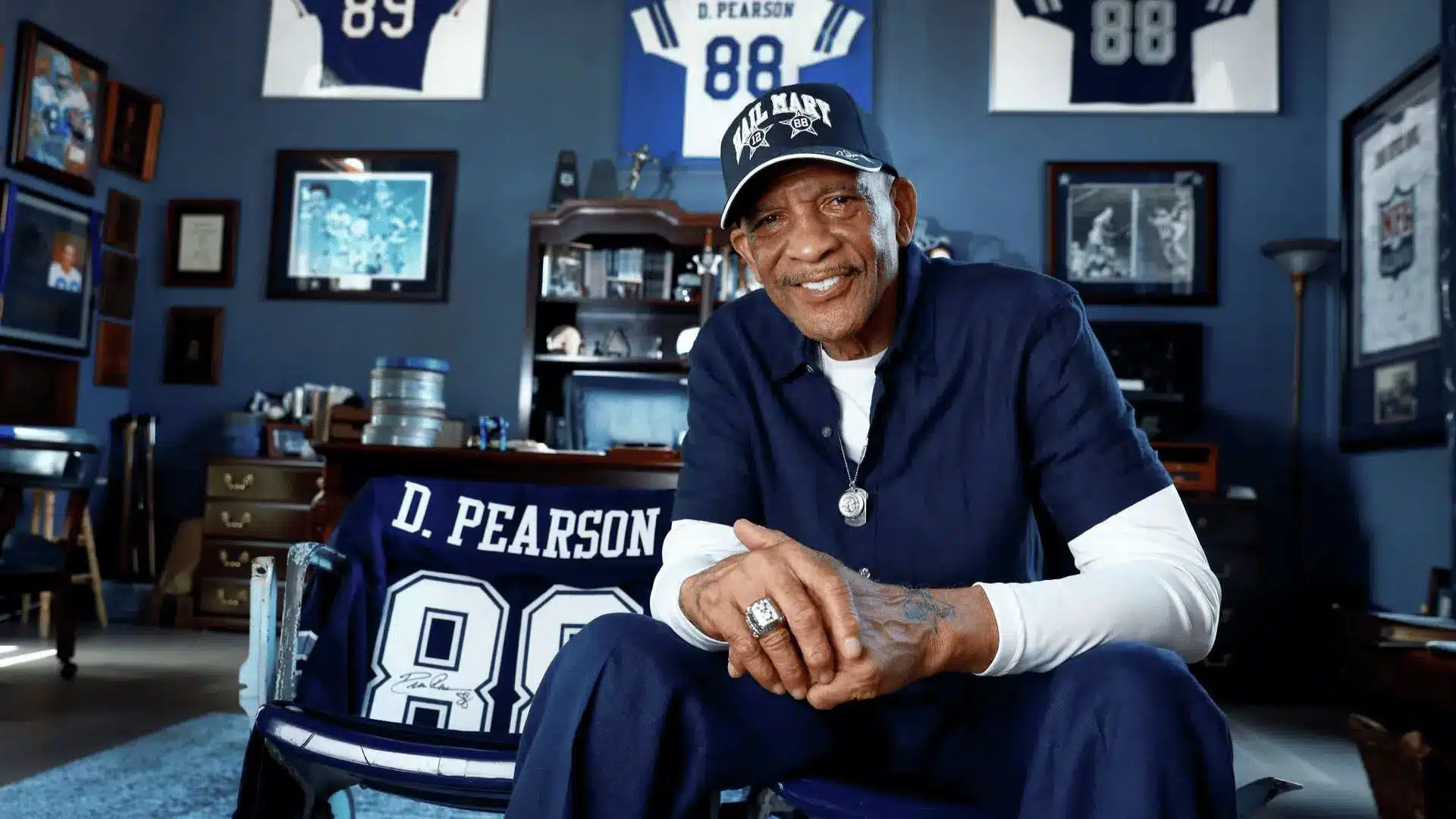 older football legend sits in a trophy filled room with framed jerseys behind him and smiles
