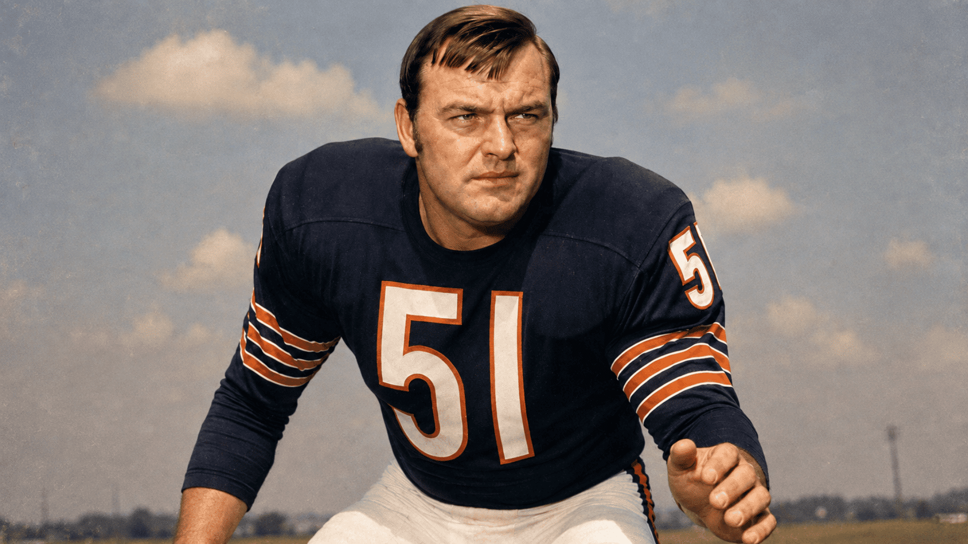 dick butkus leaning forward in a defensive football stance on the field wearing a dark chicago bears jersey