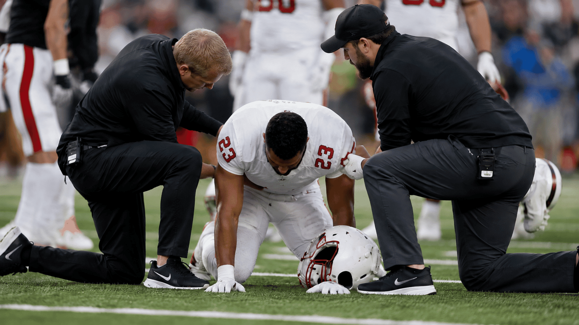 football player kneeling on the field with both hands on the turf as two trainers check on him