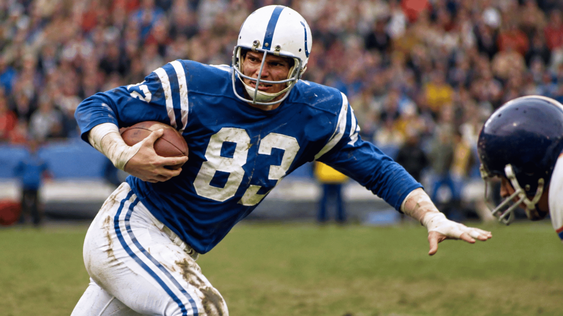 ted hendricks in a blue uniform running with the ball while reaching past a defender